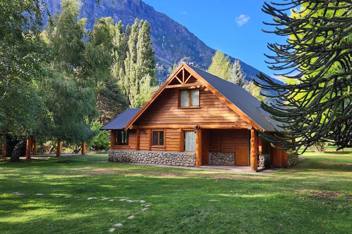 A log cabin with a stone foundation, surrounded by lush greenery and mountains in the background.