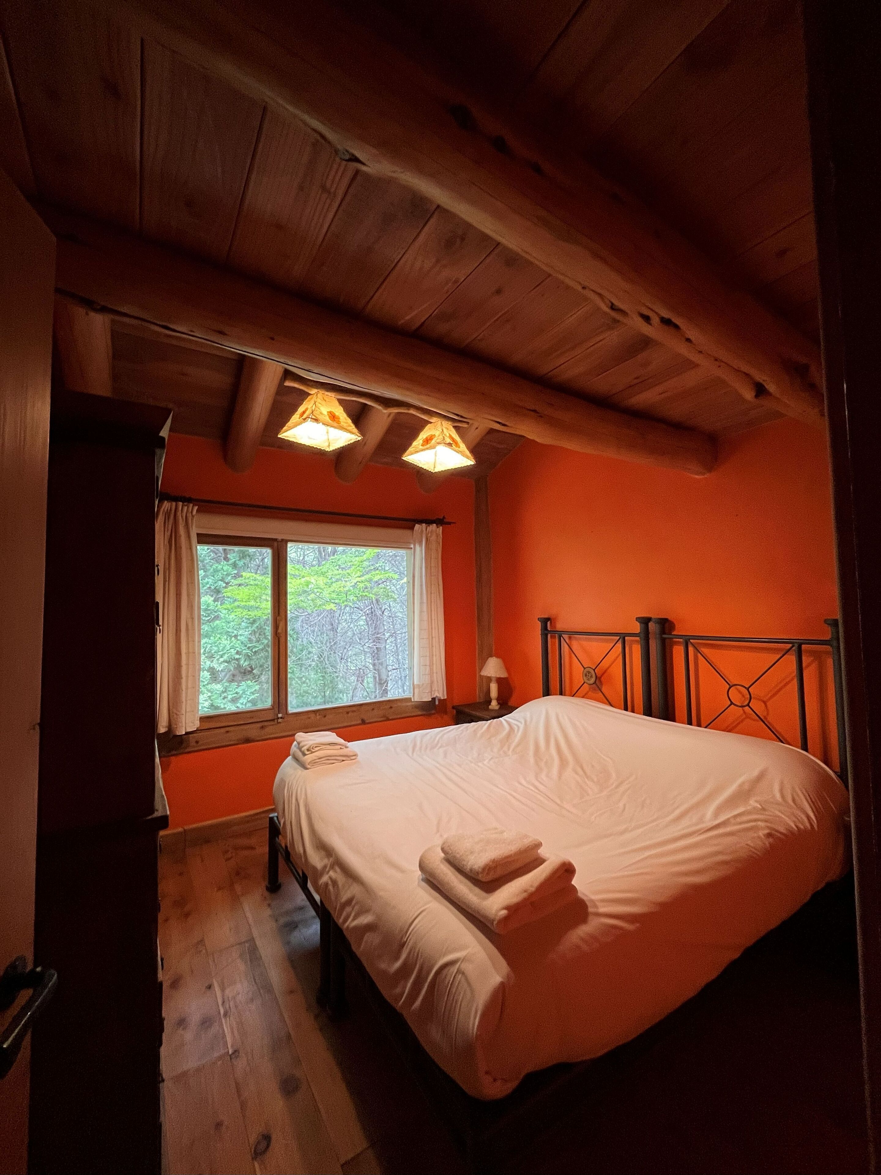 Une chambre avec un plafond en bois, un lit recouvert de draps blancs, une fenêtre avec des rideaux et une table de chevet avec une lampe.