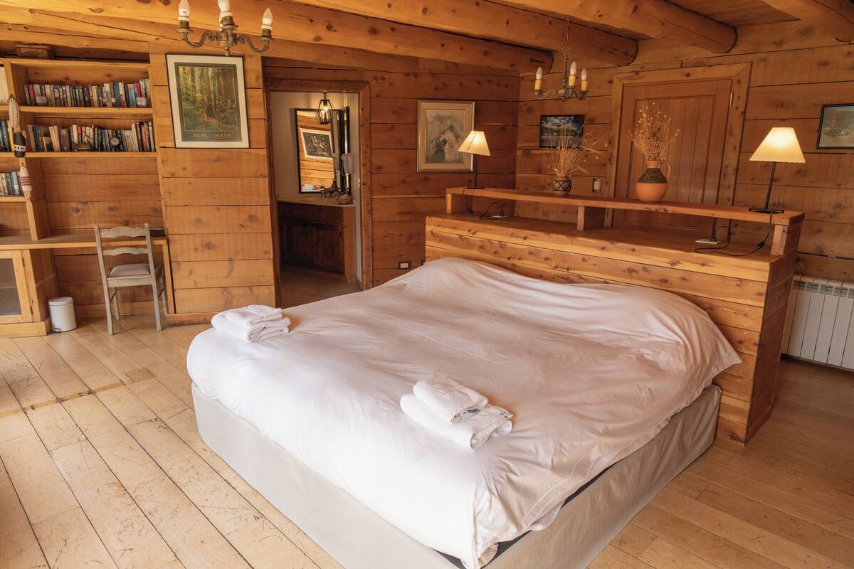 A wooden cabin bedroom with a large bed, a wooden dresser, a bookshelf, and a small desk with a chair.