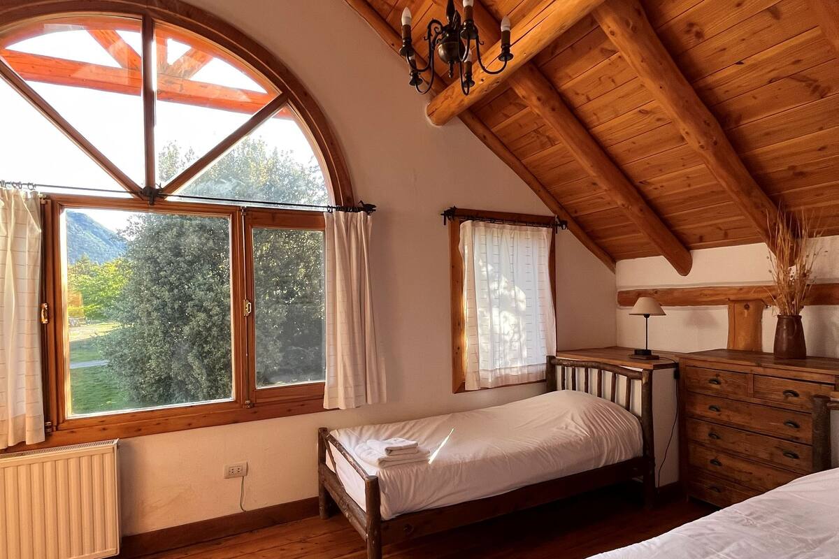 A cozy attic bedroom with wooden beams, a sloped ceiling, and a large arched window offering a view of greenery.
