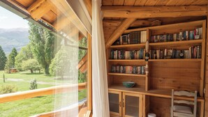 Un chalet confortable, avec un intérieur en bois, une bibliothèque remplie de livres et une vue sur un paysage verdoyant par une grande fenêtre.
