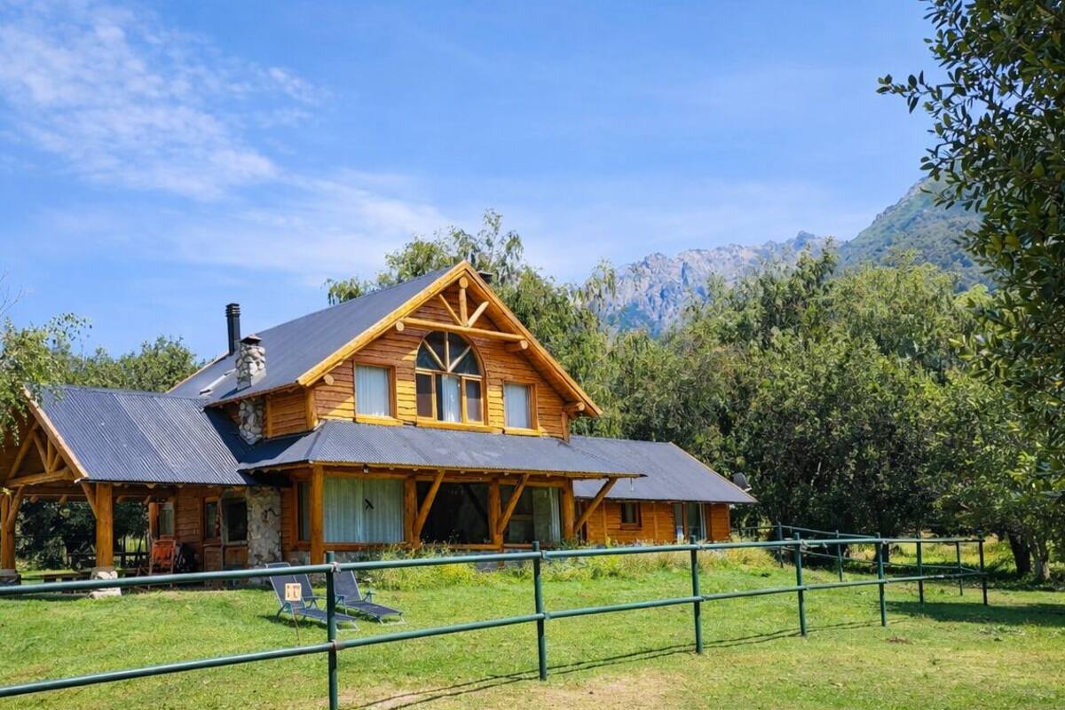A wooden cabin with a gabled roof, surrounded by a metal fence, set in a lush, green landscape with trees and mountains in the background.