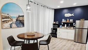Eine moderne Küche mit einem Edelstahlkühlschrank, einer Mikrowelle und einer Kaffeemaschine. Ein Essbereich mit einem runden Holztisch und vier Stühlen ist zu sehen. Ein großes Fenster mit weißen Vorhängen ist erkennbar, und eine wandmontierte Leuchte hängt über dem Tisch.