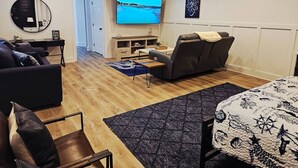 Ein Wohnzimmer mit einer Couch, einem Sessel, einem Fernseher und einem Tisch mit gemustertem Tischläufer.