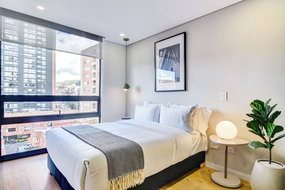 A modern bedroom with a large window, a bed with white linens, a bedside table with a lamp, and a potted plant.