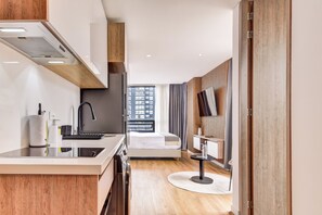 A modern hotel room with a kitchen area, a bed, a TV, and a view of the city.