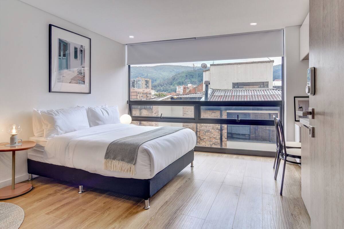 A modern bedroom with a large window, a bed, a bedside table with a lamp, and a framed picture on the wall.