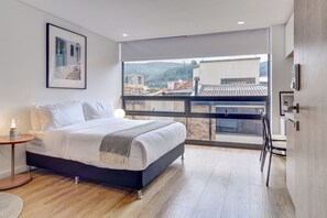 Ein modernes Schlafzimmer mit großem Fenster, einem Bett, einem Nachttisch mit Lampe und einem gerahmten Bild an der Wand.