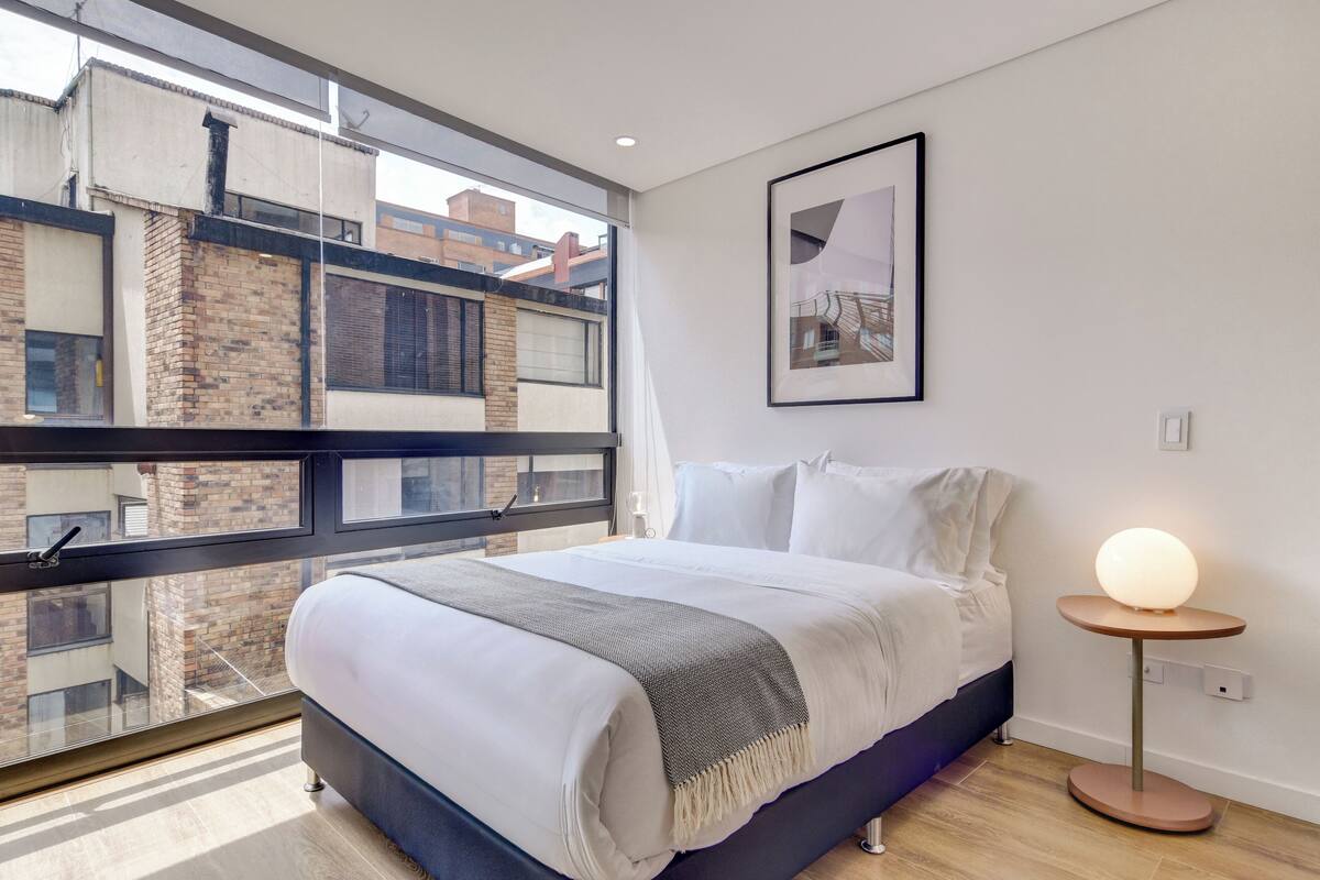 A modern bedroom with a large bed, a round table lamp, a framed picture on the wall, and a view of buildings through a large window.