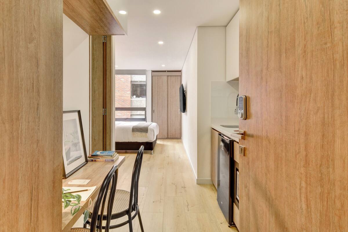 A narrow room with a bed, a desk with a chair, a small kitchen area, and a view to the outside.