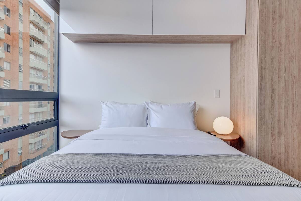 A modern hotel room with a large window, a bed with white linens, a bedside table with a lamp, and a view of a building outside.