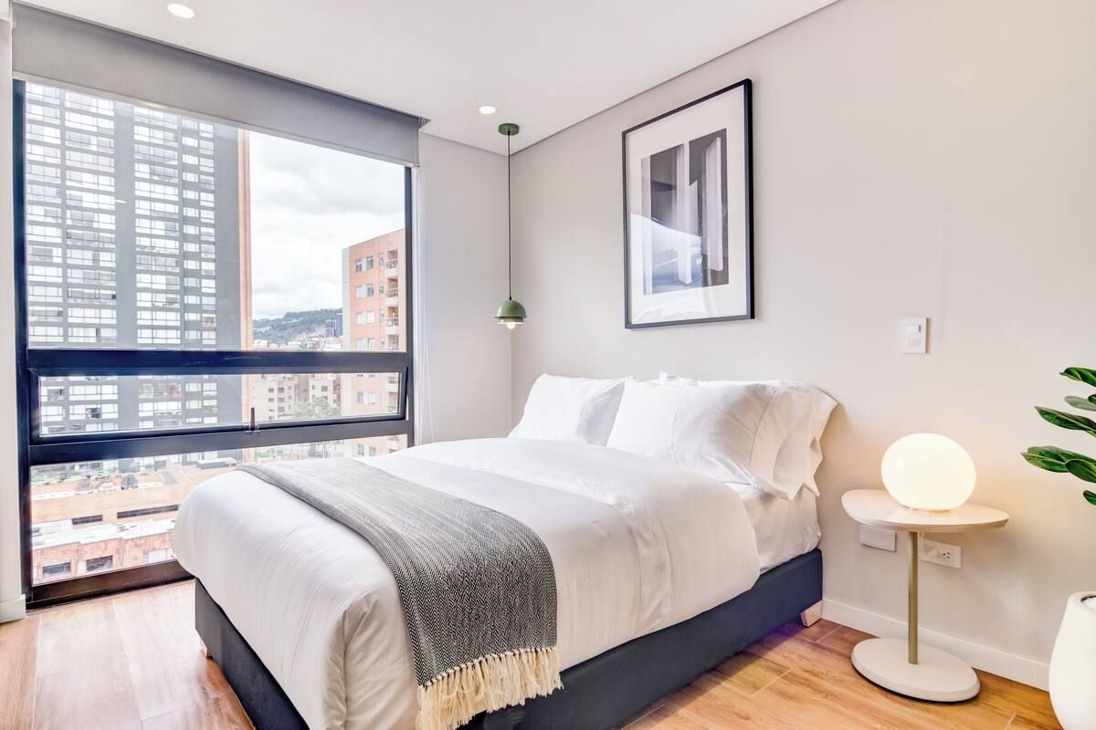 A modern bedroom with a large window, a bed with white bedding, a bedside table with a lamp, and a framed picture on the wall.