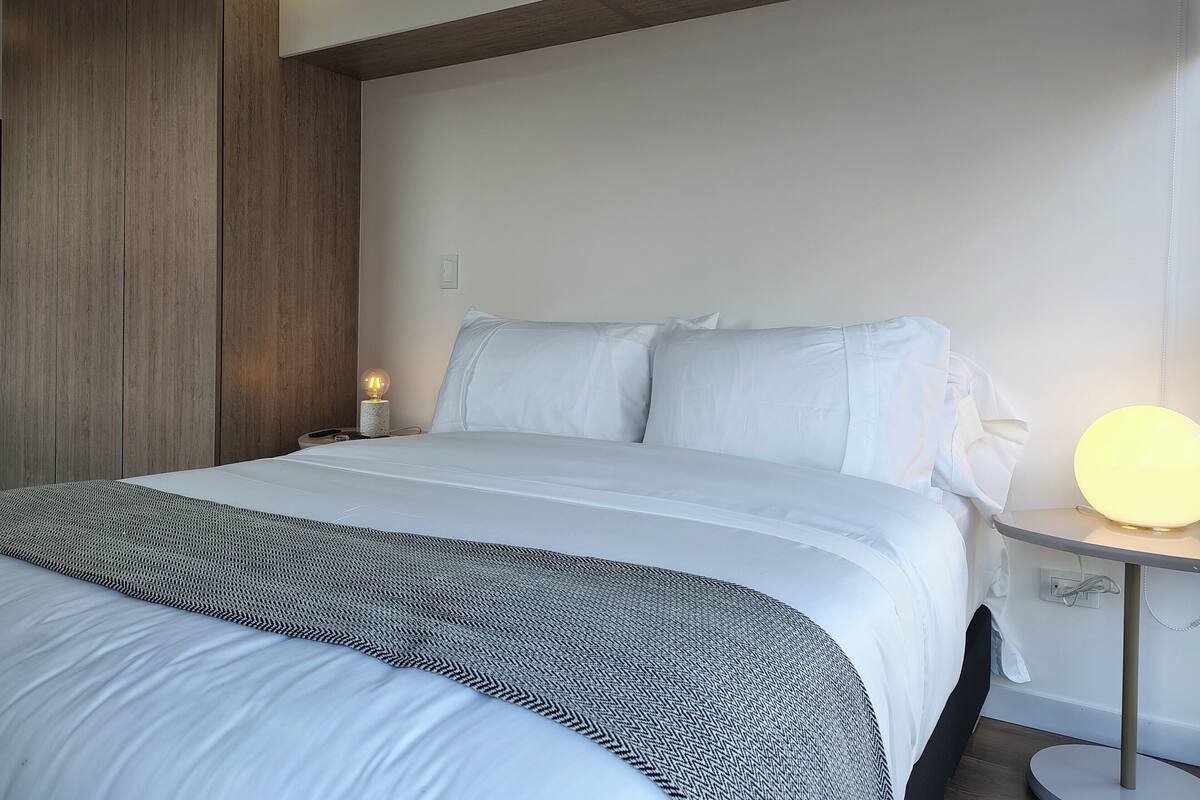 A modern hotel room with a large bed, a bedside table with a lamp, and a wooden accent wall.