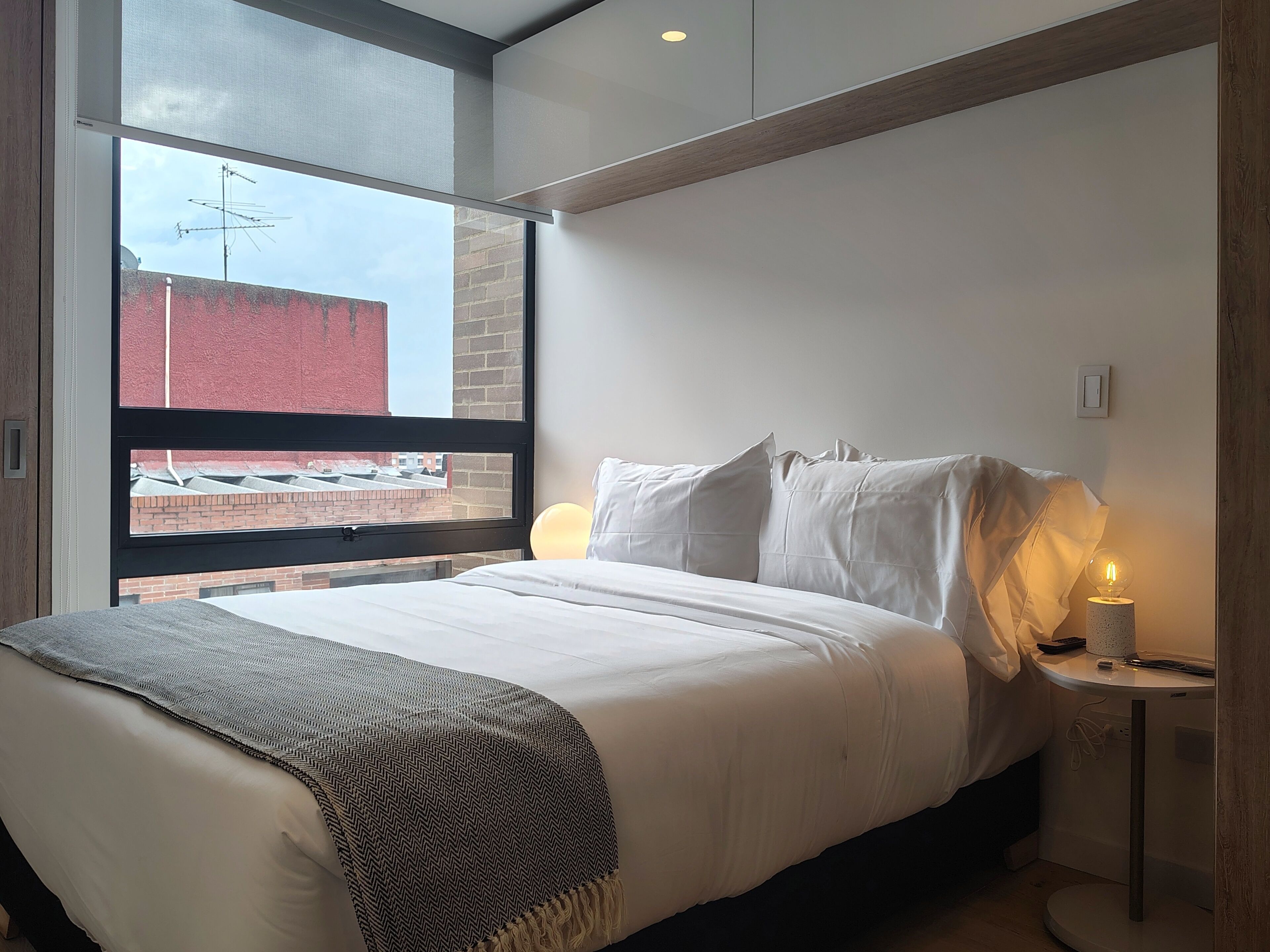 A modern bedroom with a large window, a bed with white linens, a throw blanket, and a bedside table with a lamp.