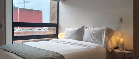 A modern bedroom with a large window, a bed with white linens, a throw blanket, and a bedside table with a lamp.