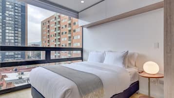 Une chambre moderne avec une grande fenêtre, un lit recouvert de draps blancs, une table de chevet avec une lampe, et une vue sur les bâtiments de la ville.