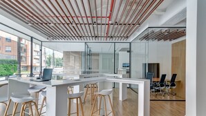 A modern office space with a white desk, bar stools, a conference table, and a large window overlooking a building. - Haus by Wynwood House (Bogotá)
