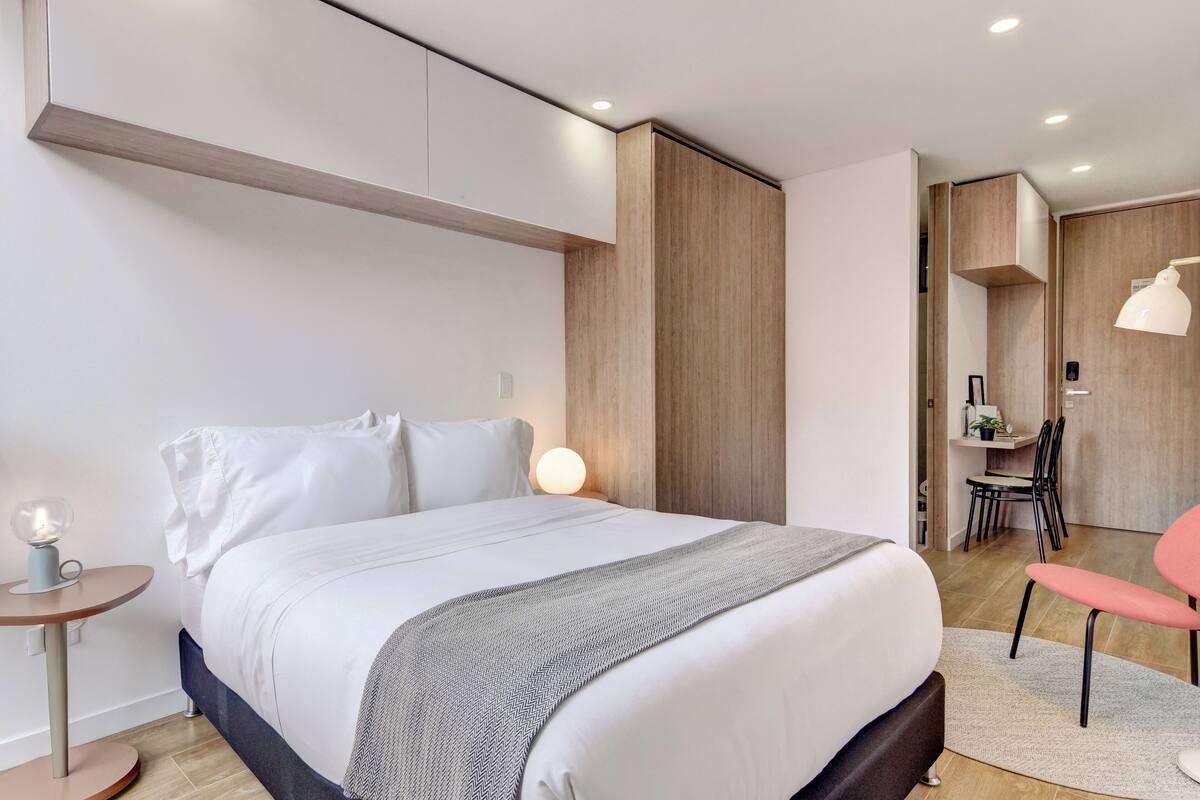 A modern hotel room with a large bed, bedside table, lamp, and a chair.