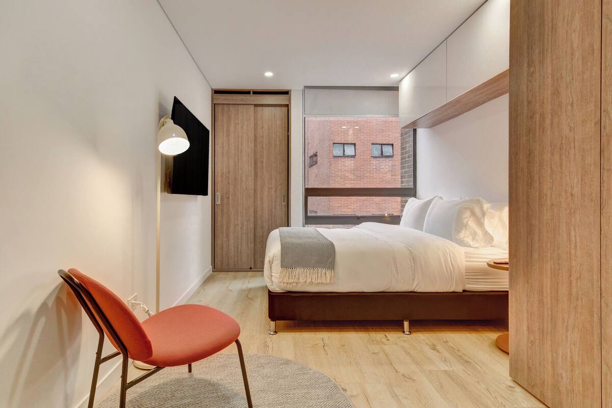 A bedroom with a bed, a chair, a desk lamp, and a window with a view of a brick building.