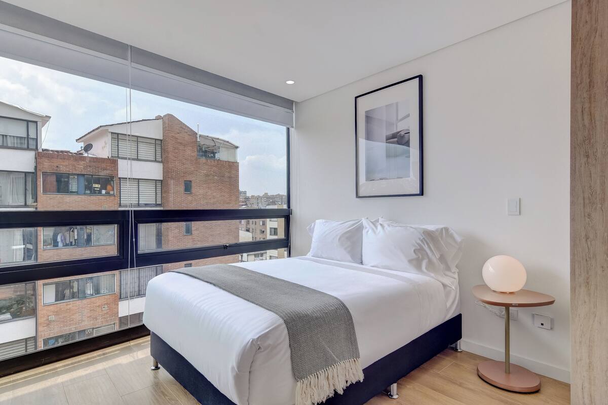 A modern bedroom with a large window, a bed with white linens, a grey throw blanket, and a round table with a light.