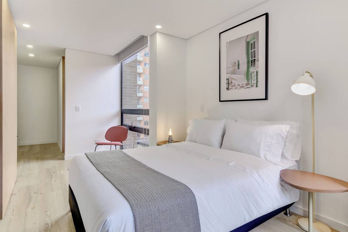 A hotel room with a bed, a desk, a chair, a framed picture on the wall, and a floor lamp.