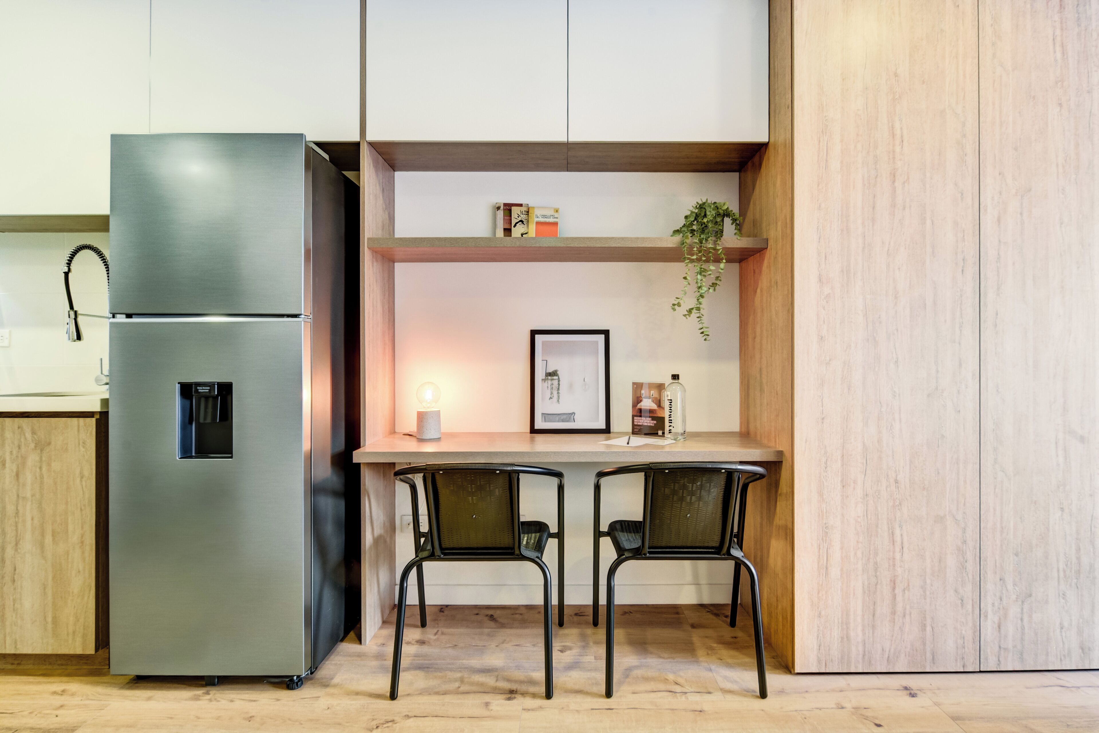 A modern kitchen with a stainless steel refrigerator, two chairs, a wooden countertop, and a shelf with a plant and framed picture.
