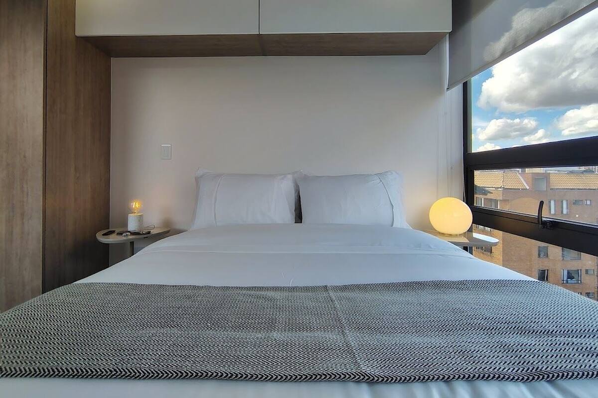 A modern hotel room with a large bed, a bedside table with a lamp, and a view of the city through a window.