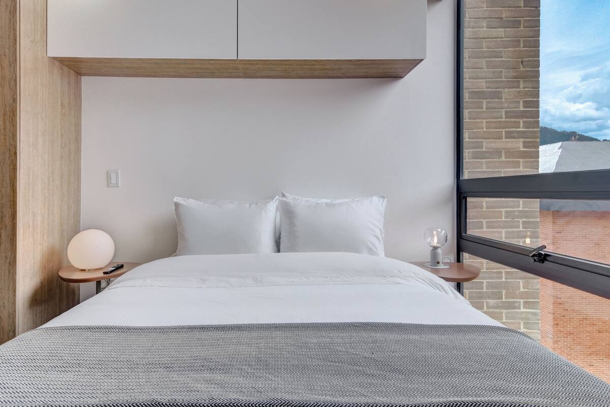 A modern hotel room with a bed, a bedside table with a lamp, a window with a view of a brick building, and a wooden headboard.