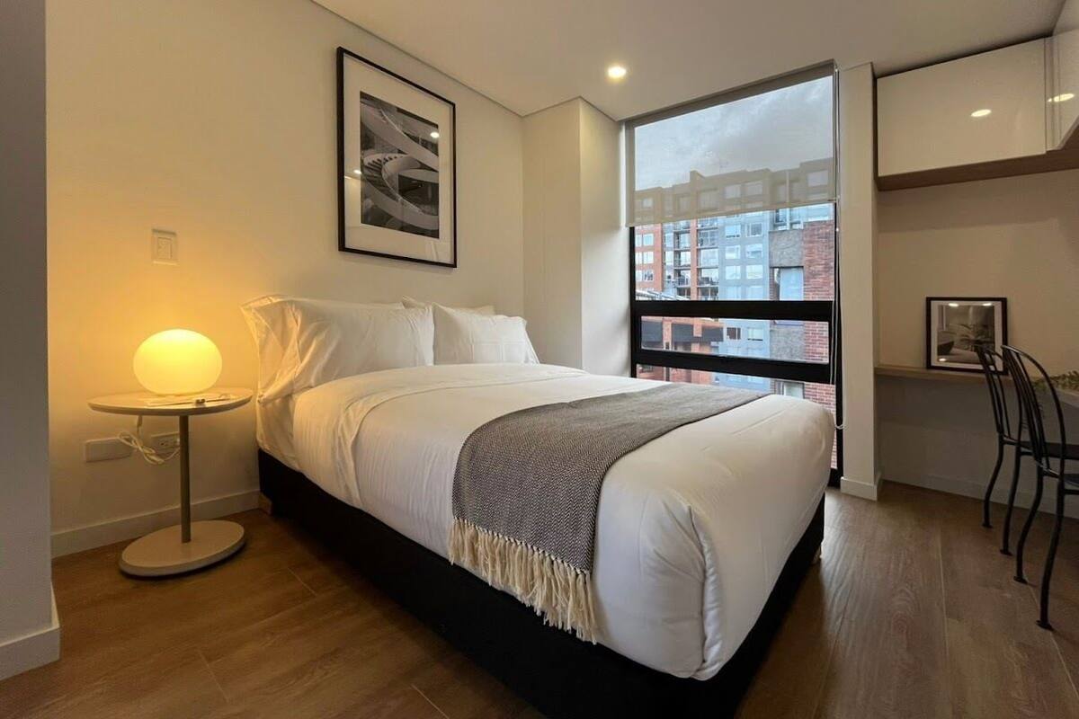 A modern hotel room with a large bed, a bedside table with a lamp, a framed picture on the wall, and a view of buildings through a window.