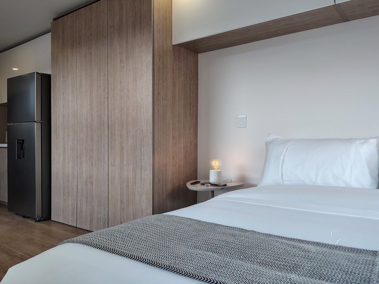 Une chambre d’hôtel comprenant un lit, une table de chevet, une lampe et un coin cuisine moderne avec un réfrigérateur.
