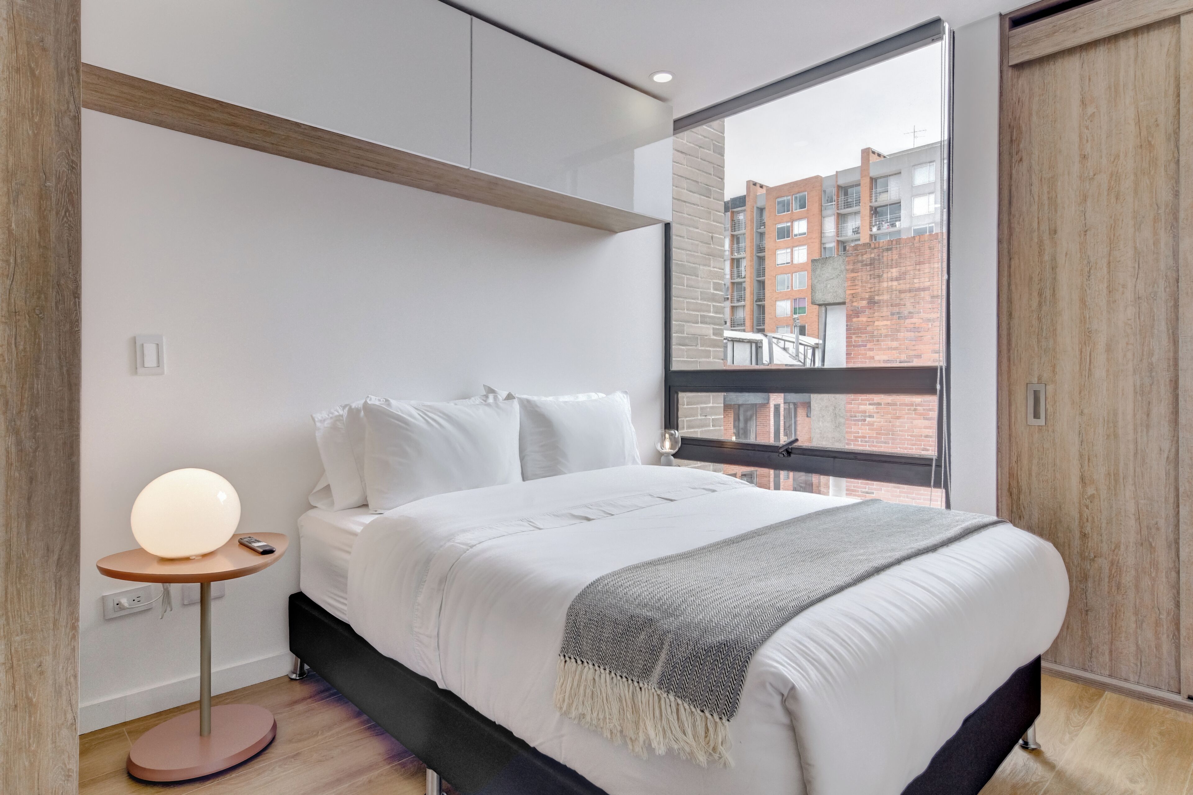 Camera da letto moderna con un letto grande, una lampada da comodino e vista su un edificio in mattoni attraverso una finestra.