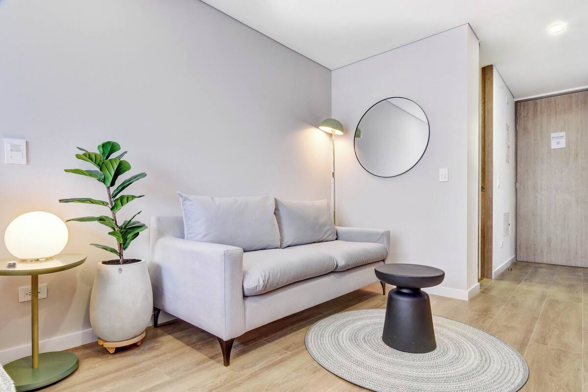 A modern living room with a sofa, a round mirror, a floor lamp, a potted plant, and a small table.