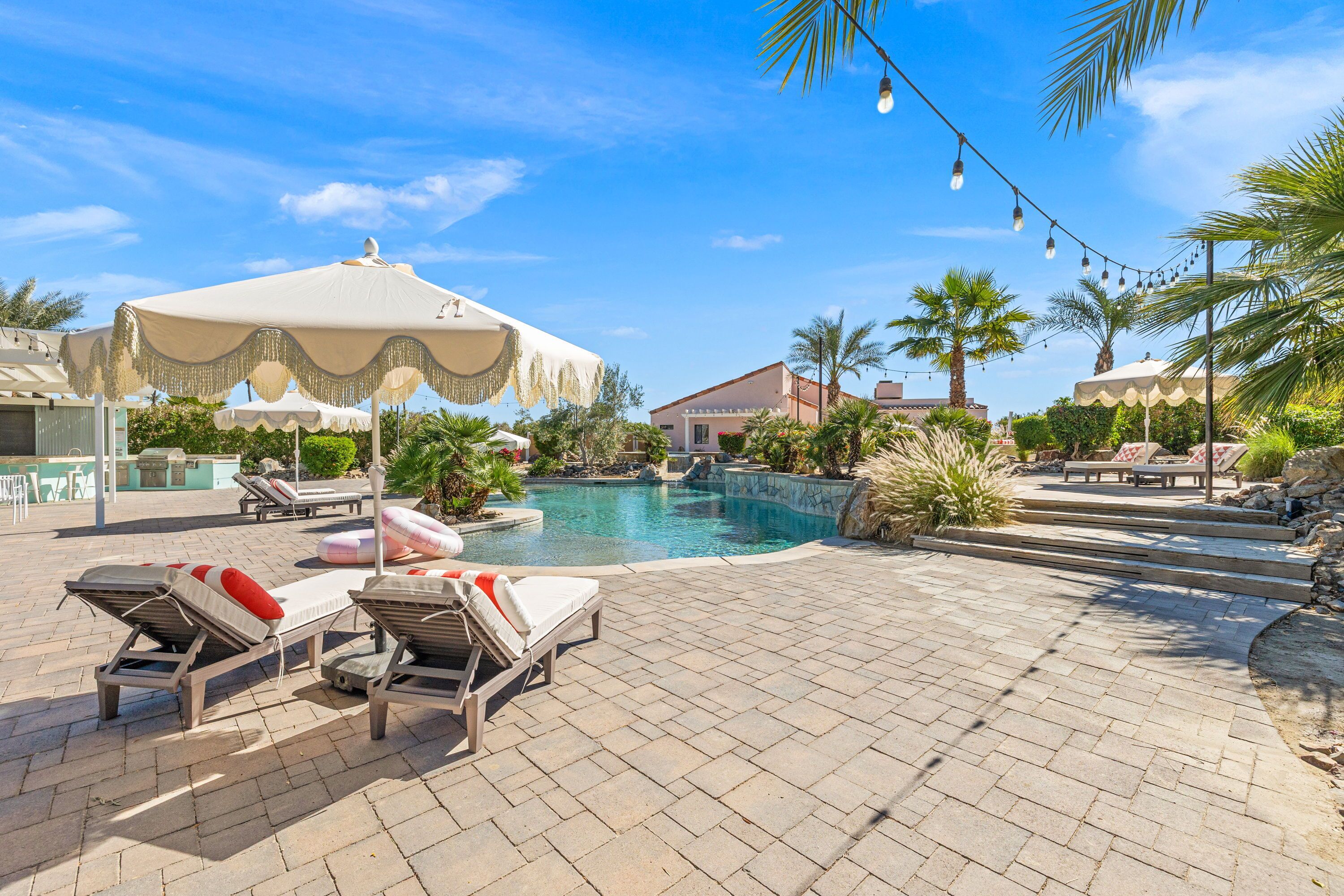 A pool area with lounge chairs and umbrellas, surrounded by palm trees and a clear blue sky.