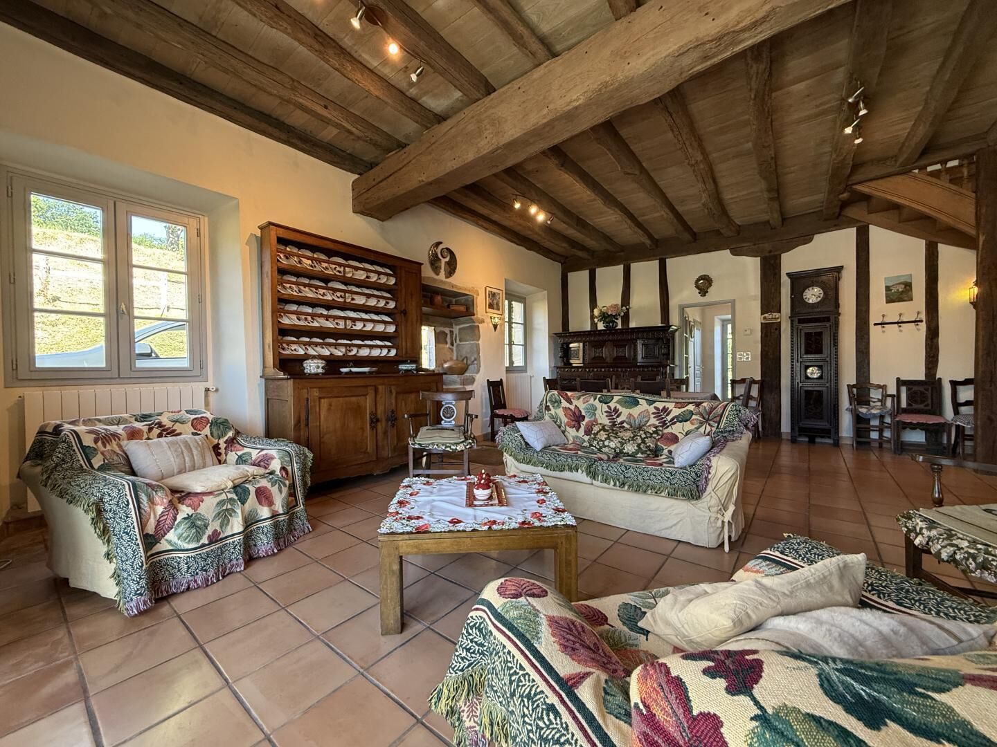 A spacious living room with exposed wooden beams, a large cabinet with dishes, and a floral-patterned sofa.