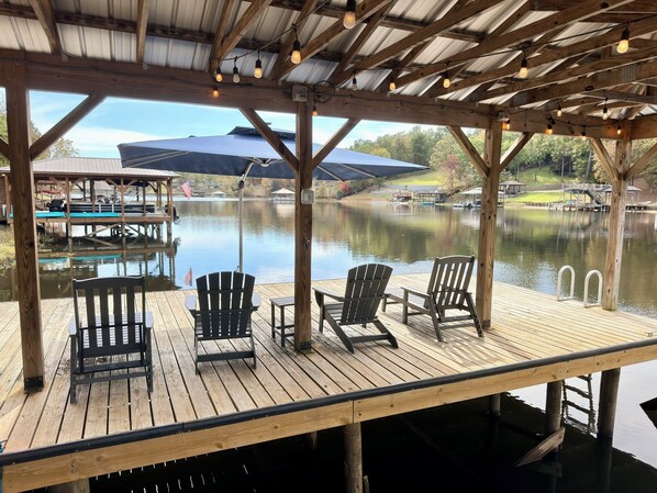 Una terraza de madera con sillas Adirondack con vistas a un lago tranquilo, un área cubierta con luces colgantes y un muelle con un barco pequeño.