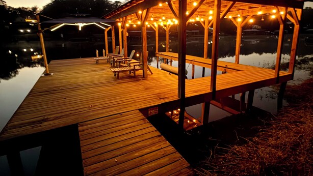 Un pabellón de madera con luces colgantes, una terraza de madera y un lago tranquilo al anochecer.