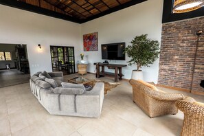 A spacious living area with a large sofa, a wicker chair, a flat-screen TV, and a potted plant. - Sahara Lodge: Nature, Design & Arabian Horses (El retiro)