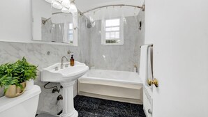 A bathroom with a pedestal sink, a bathtub, a shower with a glass door, a large mirror with lights, and a potted plant. - Spacious 3BR Luxe Escape near NYC (Yonkers)