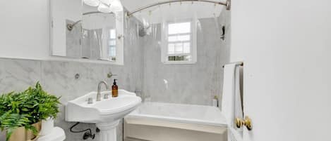 A bathroom with a pedestal sink, a bathtub, a shower with a glass door, a large mirror with lights, and a potted plant.