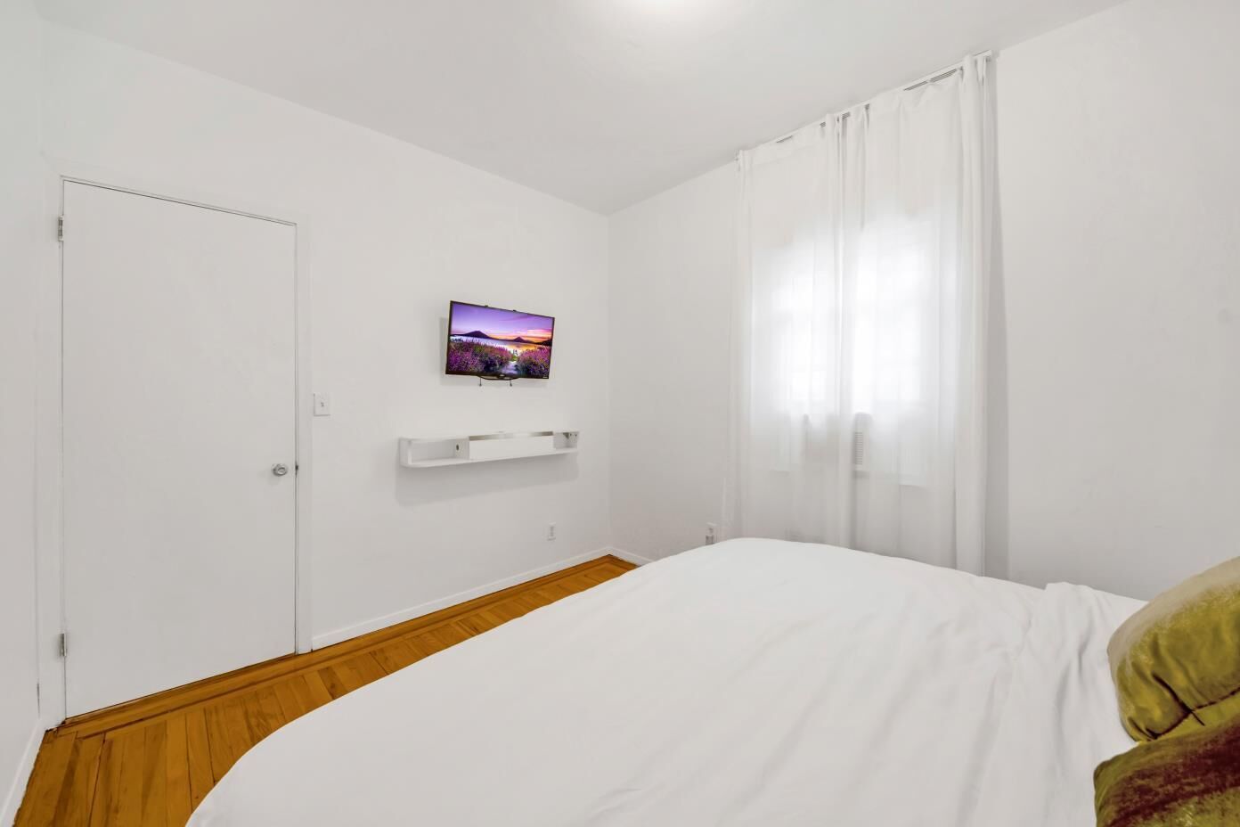 A hotel room with a bed, a mounted TV, a white shelf, and a window with sheer curtains.