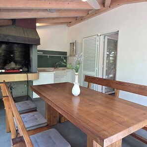 A wooden dining table with benches, a white vase with flowers, and a kitchen area with a grill and cabinets. - Holiday home with air conditioning and barbecue terrace (Baška)
