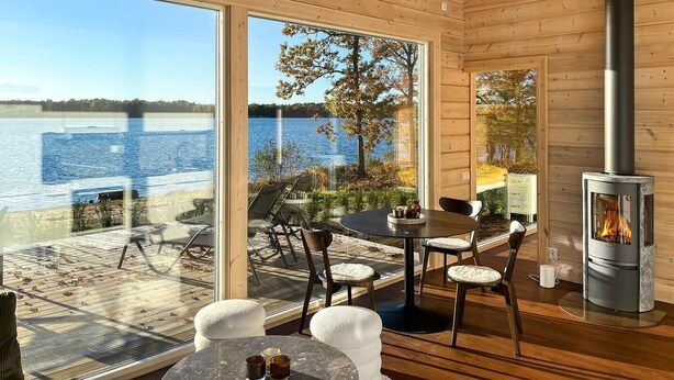 A wooden cabin with a large glass door leading to a deck overlooking a lake. Inside, there is a round table with chairs and a fireplace. - Gorgeous home in Bolmsö with lake view (Bolmsö)