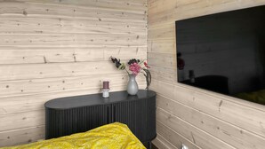 A modern bedroom with a wooden wall, a large TV, a dark console table with a vase and candle, and a yellow patterned bedspread. - Gorgeous home in Bolmsö with lake view (Bolmsö)