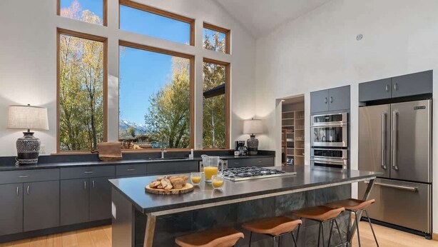 Una cocina moderna con una isla grande, taburetes de bar y vistas a árboles a través de amplias ventanas.