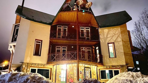 Un edificio de madera de varios pisos, con balcones, cimientos de piedra y un terreno cubierto de nieve.