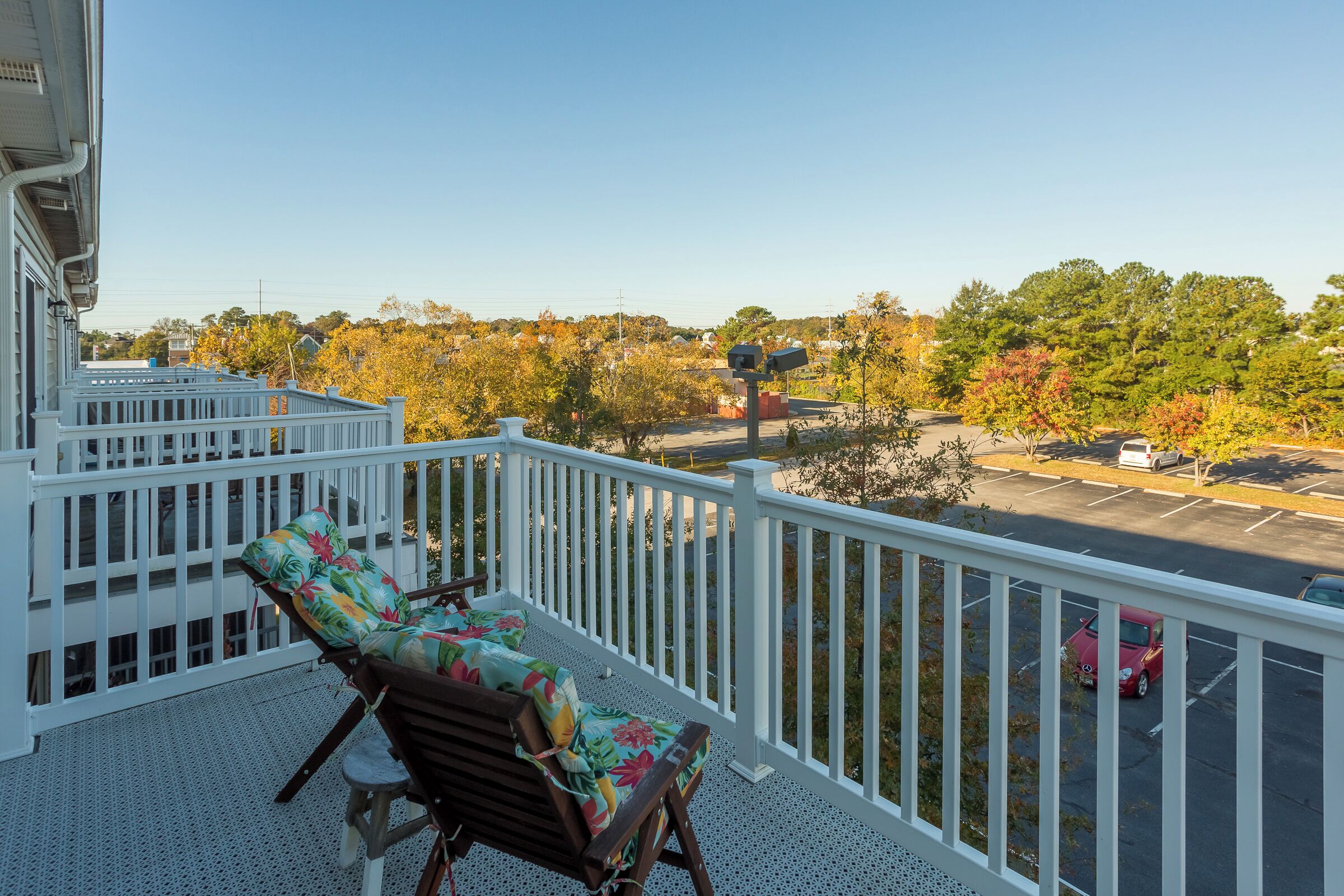 A balcony with two wooden chairs and a table, overlooking a parking lot and autumn trees.