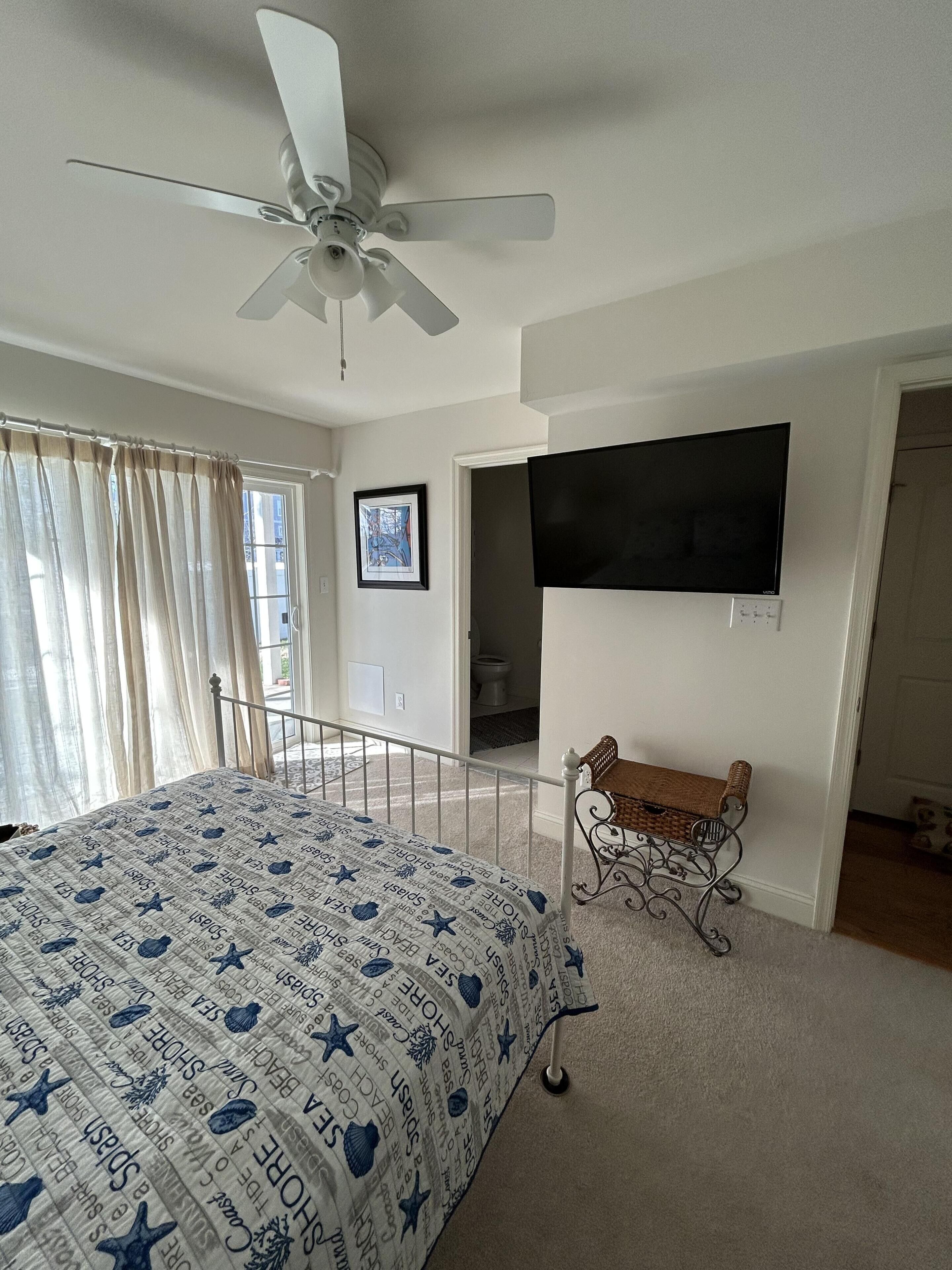 A bedroom with a bed, a ceiling fan, a TV, a window with curtains, and a door leading to another room.