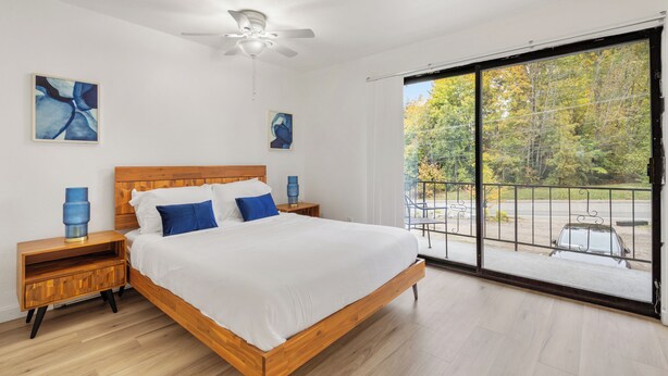 Een slaapkamer met een houten bed, witte beddengoed, een plafondventilator en een groot raam met uitzicht op bomen en een auto.