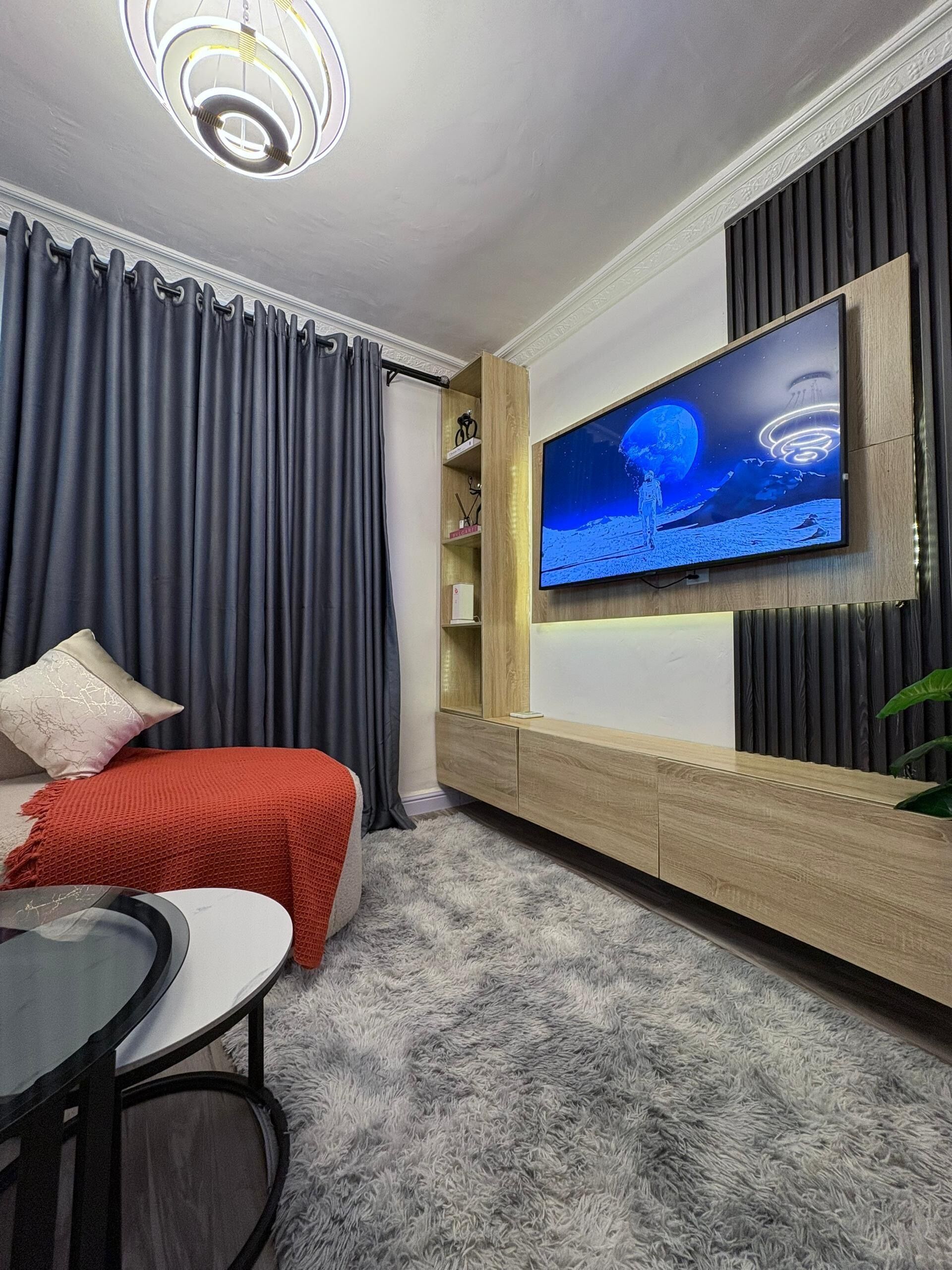 A modern living room with a large flat-screen TV mounted on a wooden wall, a cozy seating area with a red blanket, and a fluffy grey carpet.