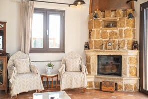 A cozy living room with a stone fireplace, two armchairs, a small table with a plant, and a window with curtains. - Sea views, sunshine, and serenity in Lakithra (Lakithra)